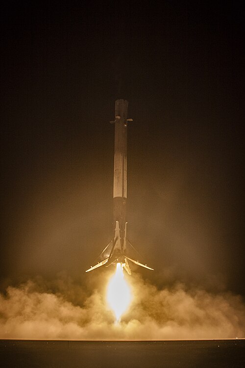 A Falcon 9 first stage descending under engine thrust to land, illuminated against a dark sky.
