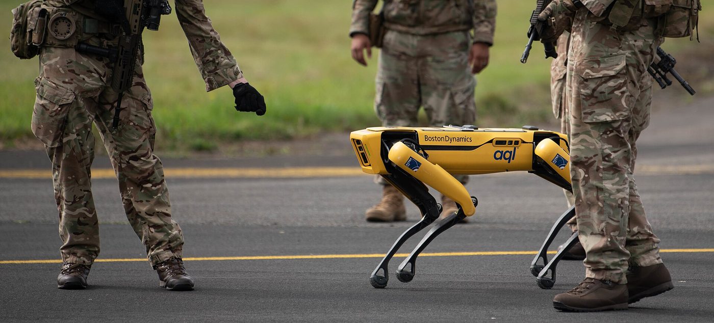 A Boston Dynamics Spot quadruped robot walking alongside personnel on a road.