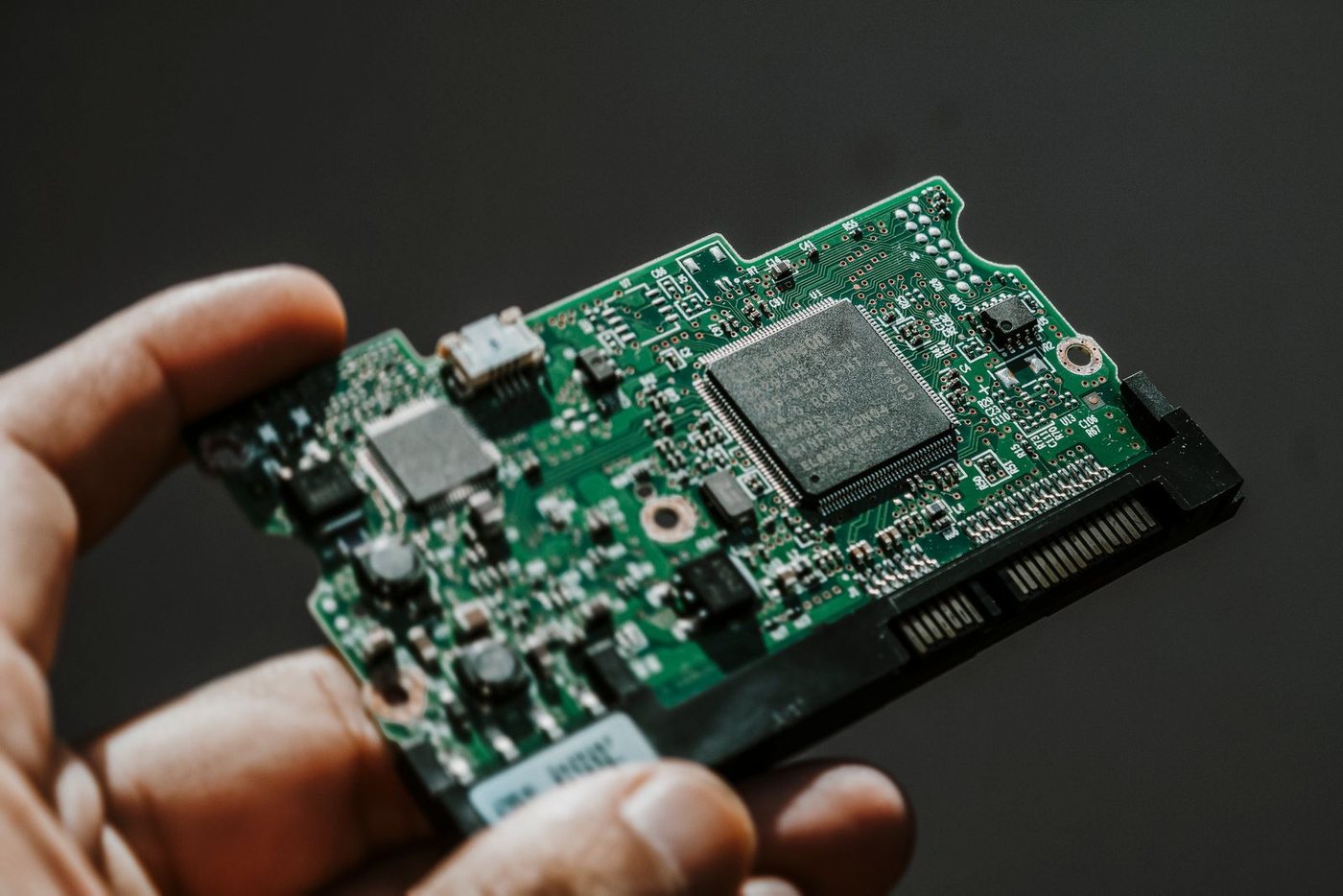 A hand holding a small populated circuit board, its processor, connectors and surface-mount components in focus.